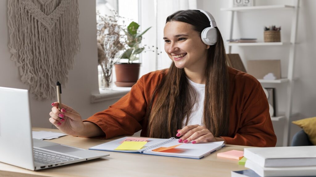 Pessoa estudando em casa com notebook, caderno e fones de ouvido.